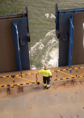 A person in a high-visibility jacket stands on the deck of a vessel, observing the water as seagulls fly over the waves. The deck is bordered by a rail with yellow and black markings, and large metal structures frame the view of the sea.