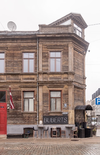A historic wooden building with a rustic appearance, featuring red-framed windows and a red door. A sign with 'BUBERTS' is displayed outside along with an assortment of mismatched chairs. A flag is hung near the door, and cobblestone pavement lies in the foreground.