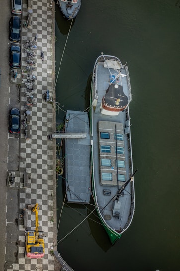 An aerial view of a docked boat in a canal, with a checkered pathway alongside. Bicycles are parked on the path and several cars are lined up on the street. A construction vehicle is visible near the bottom of the image. The boat has a white deck and several skylights.