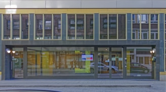 A modern, glass-fronted building with large windows reflecting the surrounding urban environment, including nearby buildings and pedestrians. The entrance features a wide set of doors with signage and colorful decals. The building's exterior is designed with a series of vertical and horizontal lines.