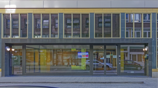 A modern, glass-fronted building with large windows reflecting the surrounding urban environment, including nearby buildings and pedestrians. The entrance features a wide set of doors with signage and colorful decals. The building's exterior is designed with a series of vertical and horizontal lines.