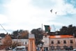 A public square with buildings, trees, and a large flagpole featuring the Algerian flag in the foreground. The sky is partly cloudy, and two birds are flying overhead. People can be seen walking around near parked vehicles.