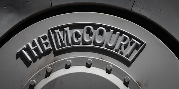 A large, metallic surface with embossed text spelling out 'THE McCOURT'. The letters are raised and cast shadows, giving a three-dimensional effect. The surface is circular, featuring bolts and rivets around the perimeter.