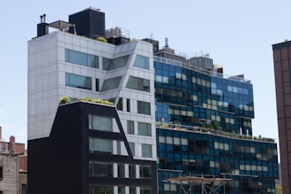 A modern urban building with a distinctive geometric design, featuring a mix of blue and white glass panels and some greenery on the rooftop terraces. The architecture blends straight lines with angled sections, and there are industrial elements like vents visible on the rooftop.