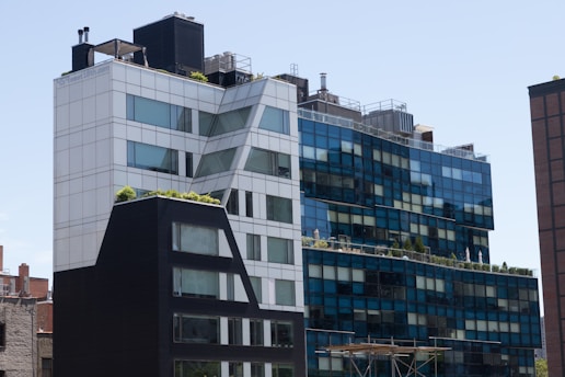 A modern urban building with a distinctive geometric design, featuring a mix of blue and white glass panels and some greenery on the rooftop terraces. The architecture blends straight lines with angled sections, and there are industrial elements like vents visible on the rooftop.