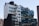 A modern urban building with a distinctive geometric design, featuring a mix of blue and white glass panels and some greenery on the rooftop terraces. The architecture blends straight lines with angled sections, and there are industrial elements like vents visible on the rooftop.