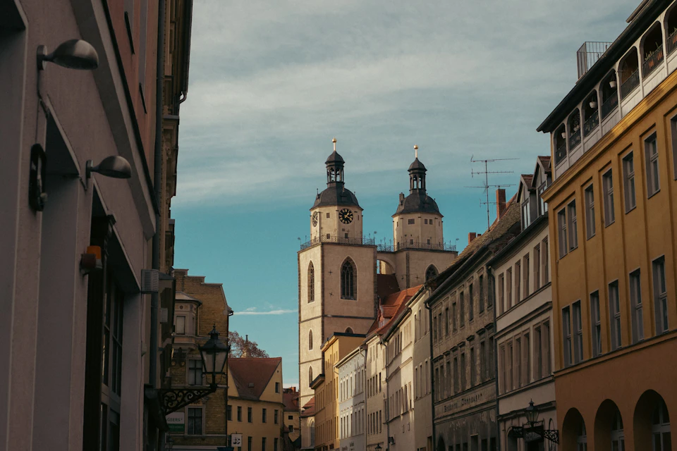 The image depicts a narrow street flanked by old, colorful buildings with white and yellow facades. The buildings feature arched windows that add to their charm. In the center of the frame is a large clock tower with two spires, standing out against the clear blue sky. A few lampposts are scattered along the street, providing illumination for pedestrians and passersby.