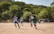 A group of young soccer players in uniform are actively running towards the ball on a dirt field. Surrounding them are lush green trees under a clear sky. The goalpost is visible in the background, with a few spectators and cars nearby.
