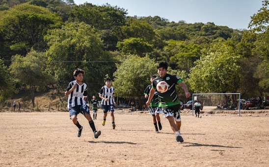 A group of young footballers in green and orange jerseys training passionately on a lush field under a bright sky.