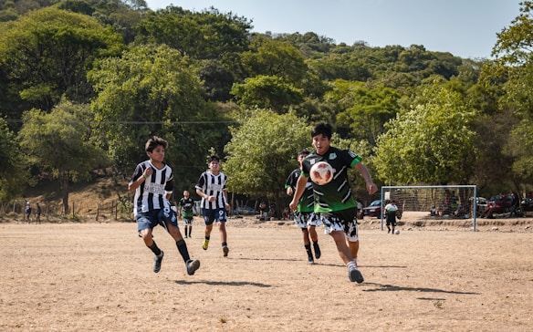 A group of young soccer players in uniform are actively running towards the ball on a dirt field. Surrounding them are lush green trees under a clear sky. The goalpost is visible in the background, with a few spectators and cars nearby.