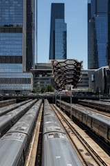 A complex urban scene featuring multiple railroad tracks with trains lined up in parallel. Above the tracks, a distinct honeycomb-like structure stands out amidst tall skyscrapers, showcasing modern architecture.