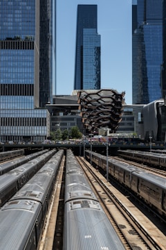 A complex urban scene featuring multiple railroad tracks with trains lined up in parallel. Above the tracks, a distinct honeycomb-like structure stands out amidst tall skyscrapers, showcasing modern architecture.