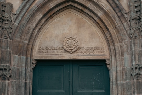 A detailed stone archway with intricate carvings and inscriptions surrounds a large, dark green wooden door. The stonework features Gothic architectural elements, and an emblem with a cross is positioned above the door.