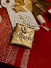 A beautifully wrapped gift rests on a red tablecloth. The box is wrapped in gold paper and adorned with a decorative string featuring small, star-shaped cutouts and bundled with white bead-like ornaments. Nearby, other wrapping materials, including a spool of ribbon and a roll of star-printed wrapping paper, are scattered, suggesting a gift-wrapping session.