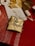 A beautifully wrapped gift rests on a red tablecloth. The box is wrapped in gold paper and adorned with a decorative string featuring small, star-shaped cutouts and bundled with white bead-like ornaments. Nearby, other wrapping materials, including a spool of ribbon and a roll of star-printed wrapping paper, are scattered, suggesting a gift-wrapping session.