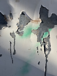 Paint peeling from an old, textured wall displaying hues of gray, brown, and green. Light shadows play across the surface, enhancing the textured appearance.
