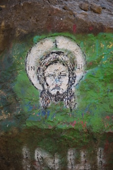 A religious painting on a rocky surface features a haloed figure with closed eyes and a somber expression. The artwork is predominantly composed of green and earthy tones, interspersed with the image of a saintly or divine figure enclosed in a circular halo.