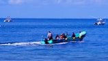 A small turquoise boat is speeding across the ocean, carrying a group of people. The occupants appear engaged in a variety of activities, possibly enjoying a leisure trip. The water is calm, and in the background, two other boats are visible; one is further away with a few people evident on deck.