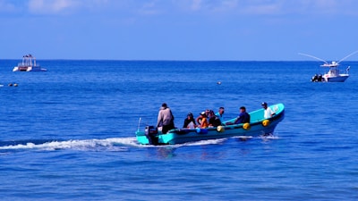 A small turquoise boat is speeding across the ocean, carrying a group of people. The occupants appear engaged in a variety of activities, possibly enjoying a leisure trip. The water is calm, and in the background, two other boats are visible; one is further away with a few people evident on deck.