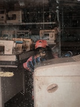 A person wearing a red cap and colorful jacket is inside a room filled with sewing machines. The scene appears to be viewed through a window with water droplets, adding a textured effect. The person seems to be engaged in an activity, possibly related to sewing or using the equipment present.