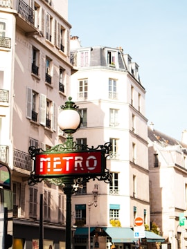 An ornate metro sign in a classic style stands prominently in front of an elegant building with cream-colored walls and traditional Parisian architecture. The building features numerous windows with decorative railings, characteristic of the city's aesthetic, under a clear blue sky.