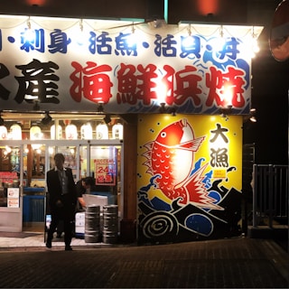 The image portrays the exterior of a brightly lit Japanese seafood restaurant at night. The signage is prominent with Japanese characters in blue and red, and a large, colorful image of a red fish against a yellow background, surrounded by waves. Lanterns with additional Japanese writing hang above the entrance. There are two people visible; one appears to be walking out of the establishment, and another is inside, engaged in some task.
