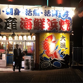 The image portrays the exterior of a brightly lit Japanese seafood restaurant at night. The signage is prominent with Japanese characters in blue and red, and a large, colorful image of a red fish against a yellow background, surrounded by waves. Lanterns with additional Japanese writing hang above the entrance. There are two people visible; one appears to be walking out of the establishment, and another is inside, engaged in some task.