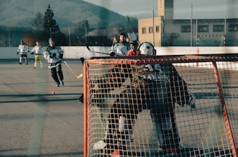 Several hockey players are engaging in a game on an outdoor rink. The scene focuses on the goalie in front of the net, with players approaching in the background, one holding a hockey stick about to make a play.