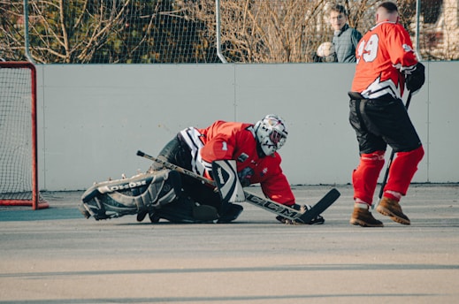 Two hockey players wearing red jerseys and black gear are on an outdoor rink. One player is kneeling on the ground with a hockey stick, appearing to block a shot, while the other skates nearby. In the background, there is a goal post and someone observing the game.