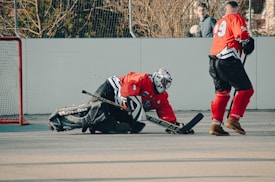 Two hockey players wearing red jerseys and black gear are on an outdoor rink. One player is kneeling on the ground with a hockey stick, appearing to block a shot, while the other skates nearby. In the background, there is a goal post and someone observing the game.