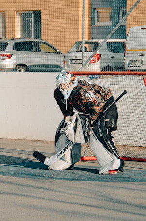 A person wearing a hockey goalie outfit is standing in front of a goal net on a concrete surface. The goalie is equipped with pads, a helmet, and gloves. In the background, there are parked cars and buildings, and a netting provides a barrier.