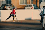 A person wearing a red jersey and cap is playing street hockey. They are focused on hitting a small red ball with a hockey stick on an outdoor court. Another person in a green and white jersey is visible, and a spectator sits nearby on a bench. Cars are parked behind a wire fence, and buildings with windows are in the background.