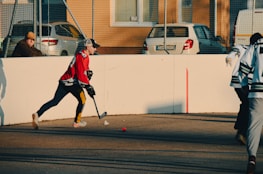 A person wearing a red jersey and cap is playing street hockey. They are focused on hitting a small red ball with a hockey stick on an outdoor court. Another person in a green and white jersey is visible, and a spectator sits nearby on a bench. Cars are parked behind a wire fence, and buildings with windows are in the background.