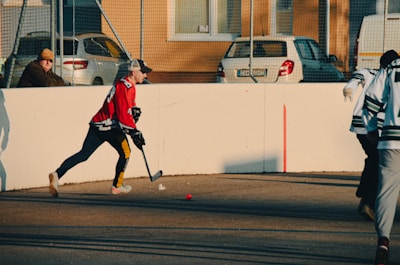 A person wearing a red jersey and cap is playing street hockey. They are focused on hitting a small red ball with a hockey stick on an outdoor court. Another person in a green and white jersey is visible, and a spectator sits nearby on a bench. Cars are parked behind a wire fence, and buildings with windows are in the background.