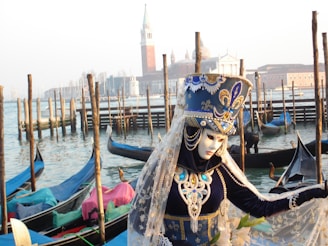 A person wearing an elaborately decorated costume and mask stands near a canal with gondolas in the background. The costume is ornate, featuring intricate patterns in blue, white, and gold, with a high headpiece adorned with jewels. In the background, traditional wooden poles rise from the water, and a classic Venetian skyline with a tower and church domes is visible across the canal.