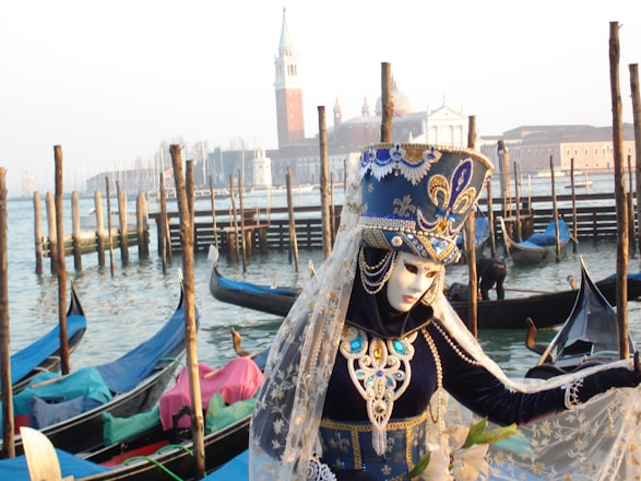 A person wearing an elaborately decorated costume and mask stands near a canal with gondolas in the background. The costume is ornate, featuring intricate patterns in blue, white, and gold, with a high headpiece adorned with jewels. In the background, traditional wooden poles rise from the water, and a classic Venetian skyline with a tower and church domes is visible across the canal.
