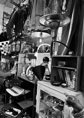 A vintage store display featuring a variety of items including jackets hanging from a pipe, several table lamps, books on a small shelf, and a glass case labeled 'VENDOR #39' containing trinkets and souvenirs. An accordion is visible on a lower shelf, and a variety of bric-a-brac and decorative items clutter the shelves. The setting suggests an eclectic mix of retro and antique objects.
