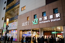 A busy urban area featuring the exterior of a building with prominent signs for 'atr&eacute; 1' and 'Akihabara Station'. People are walking in front of the building, and there are colorful advertisements and window displays visible. The structure is well-lit, suggesting it is evening or night time.