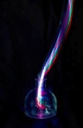 A dark background accentuates a glass container from which vibrant, multicolored light trails emerge, creating a dynamic, energetic visual effect.