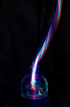 A dark background accentuates a glass container from which vibrant, multicolored light trails emerge, creating a dynamic, energetic visual effect.