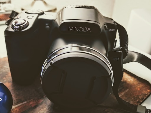 A black digital camera with the brand name displayed on the lens barrel, placed on a textured surface. The camera strap is visible on the right side, and the overall setting suggests an indoor environment with soft lighting.