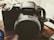 A black digital camera with the brand name displayed on the lens barrel, placed on a textured surface. The camera strap is visible on the right side, and the overall setting suggests an indoor environment with soft lighting.
