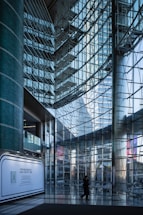 A modern interior with large glass walls reflecting light, structural metal beams creating a geometric pattern. A lone figure walks across the shiny tiled floor, and a promotional display board is positioned against a column.