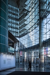 A modern interior with large glass walls reflecting light, structural metal beams creating a geometric pattern. A lone figure walks across the shiny tiled floor, and a promotional display board is positioned against a column.