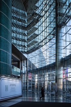 A modern interior with large glass walls reflecting light, structural metal beams creating a geometric pattern. A lone figure walks across the shiny tiled floor, and a promotional display board is positioned against a column.