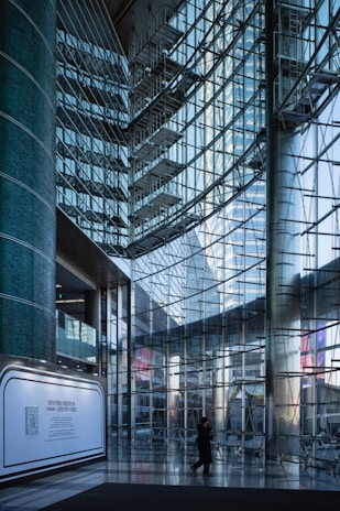 A modern interior with large glass walls reflecting light, structural metal beams creating a geometric pattern. A lone figure walks across the shiny tiled floor, and a promotional display board is positioned against a column.