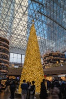 A large golden Christmas tree stands in a spacious, modern indoor setting with a high glass ceiling. The tree is adorned with numerous small lights and silver ornaments. Behind it are two tall, circular shelves filled with books, giving the appearance of a library setting. Several people are gathered around the base of the tree, some taking pictures. The environment is busy and lively.