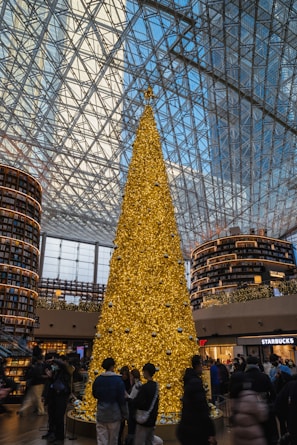 A large golden Christmas tree stands in a spacious, modern indoor setting with a high glass ceiling. The tree is adorned with numerous small lights and silver ornaments. Behind it are two tall, circular shelves filled with books, giving the appearance of a library setting. Several people are gathered around the base of the tree, some taking pictures. The environment is busy and lively.