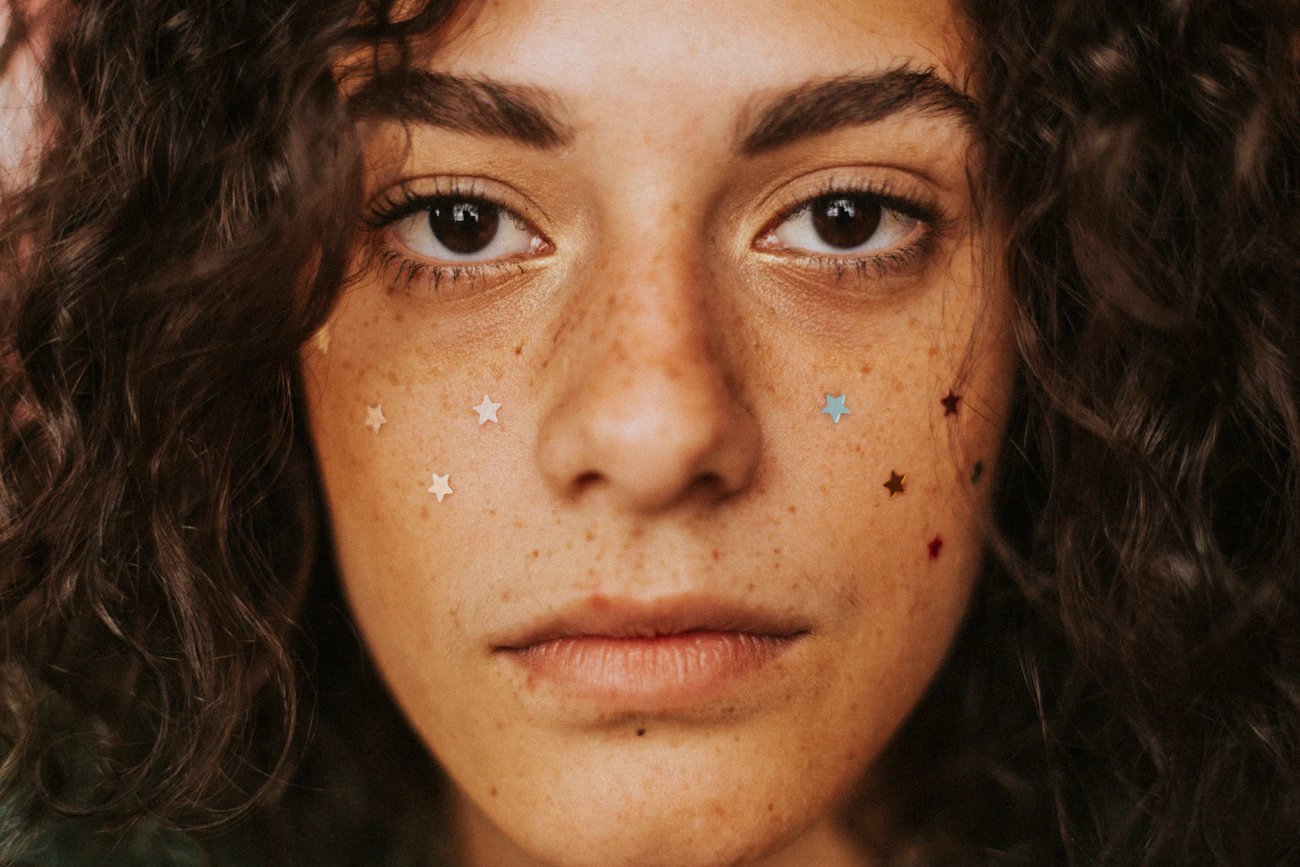 Close-up beauty portrait of a woman's face with visible natural freckles and pigmentation across the cheeks and nose, representing the pigmentation and tone concerns in this category