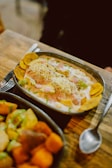A metal oval dish contains a colorful meal with chunks of green avocado, cooked chickpeas, shredded cheese, and plantain chips, topped with a creamy sauce and garnished with herbs. Another dish filled with various cooked vegetables, such as potatoes and sweet potatoes, is partially visible in the foreground. Both dishes rest on a rustic wooden table beside a spoon.